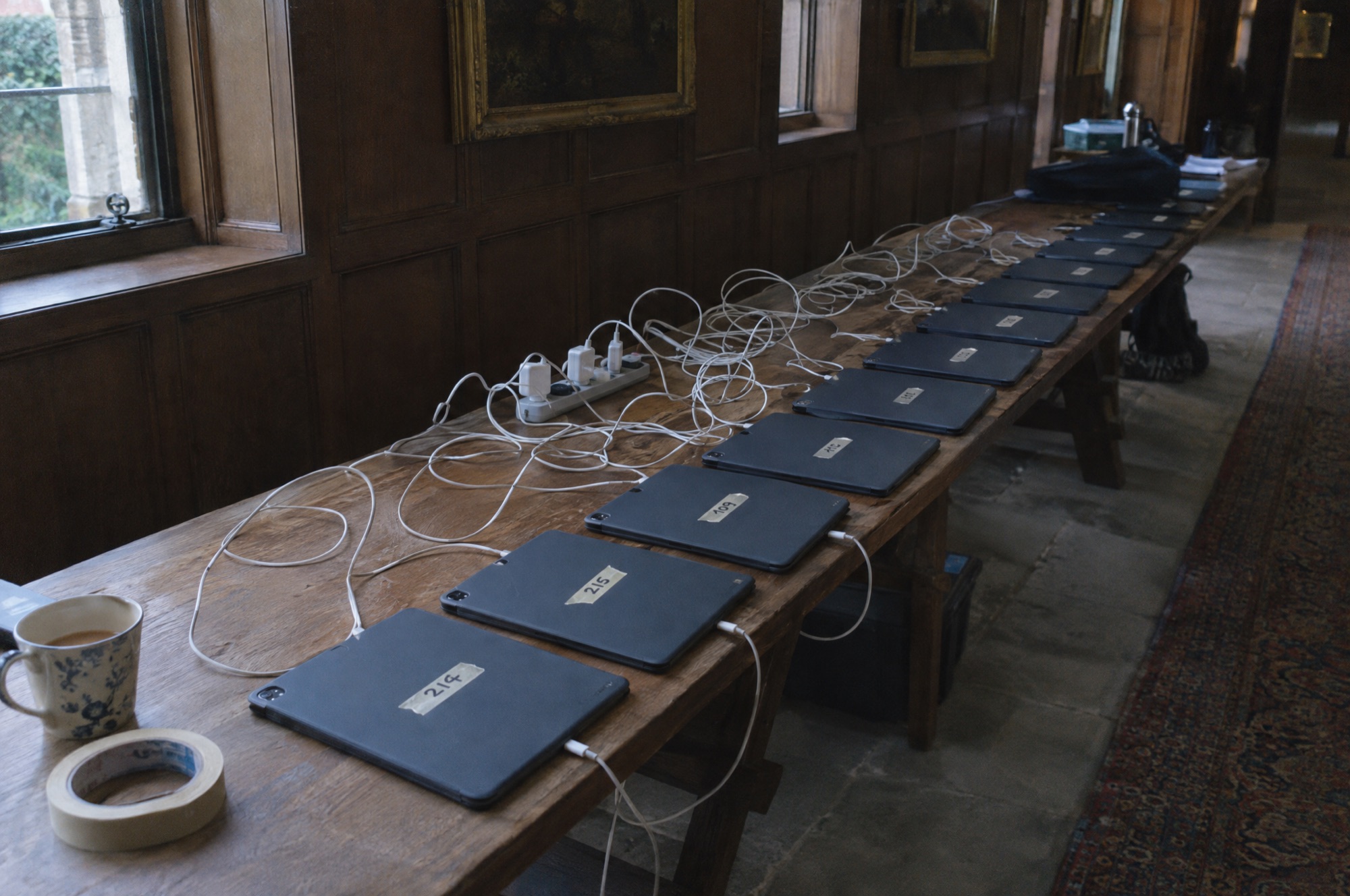 A long table of iPads on charge, each with a room number on masking tape